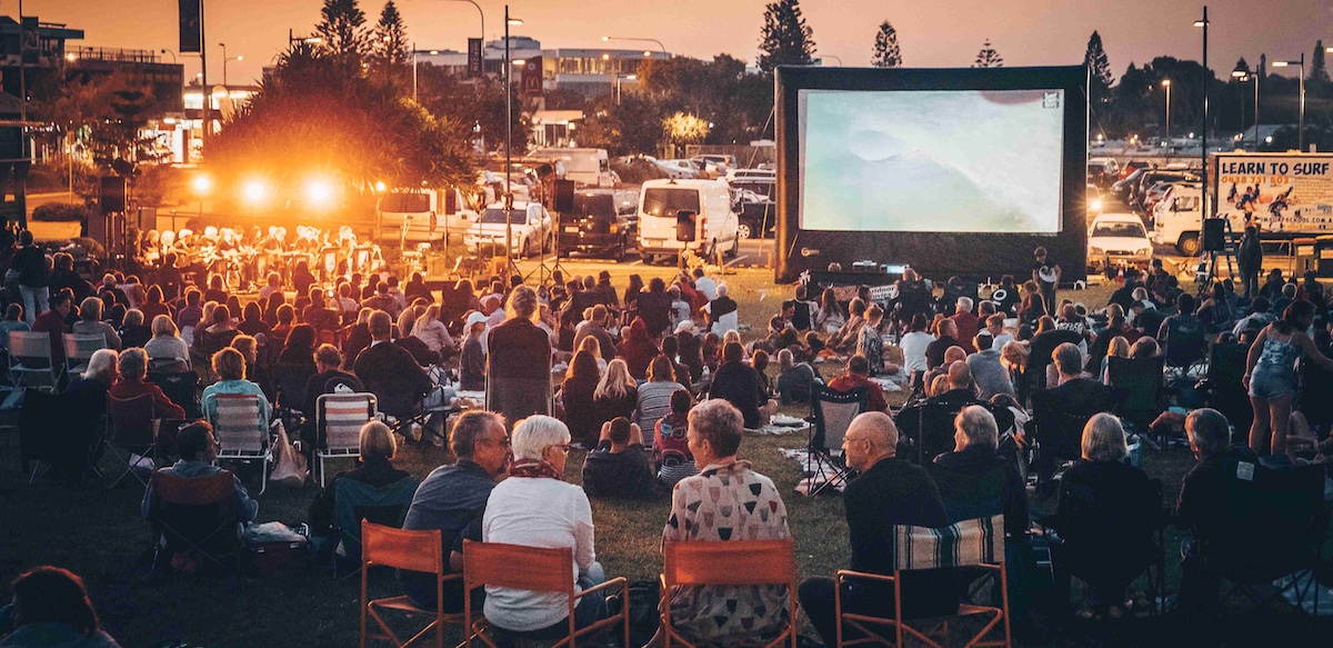 A crowd sitting under the stars in front of a large outdoor movie screen by the beach.